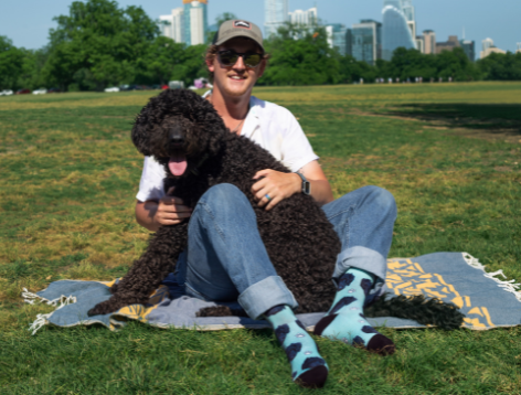 Man wearing blue dog face socks, sitting in grass, holding black dog 