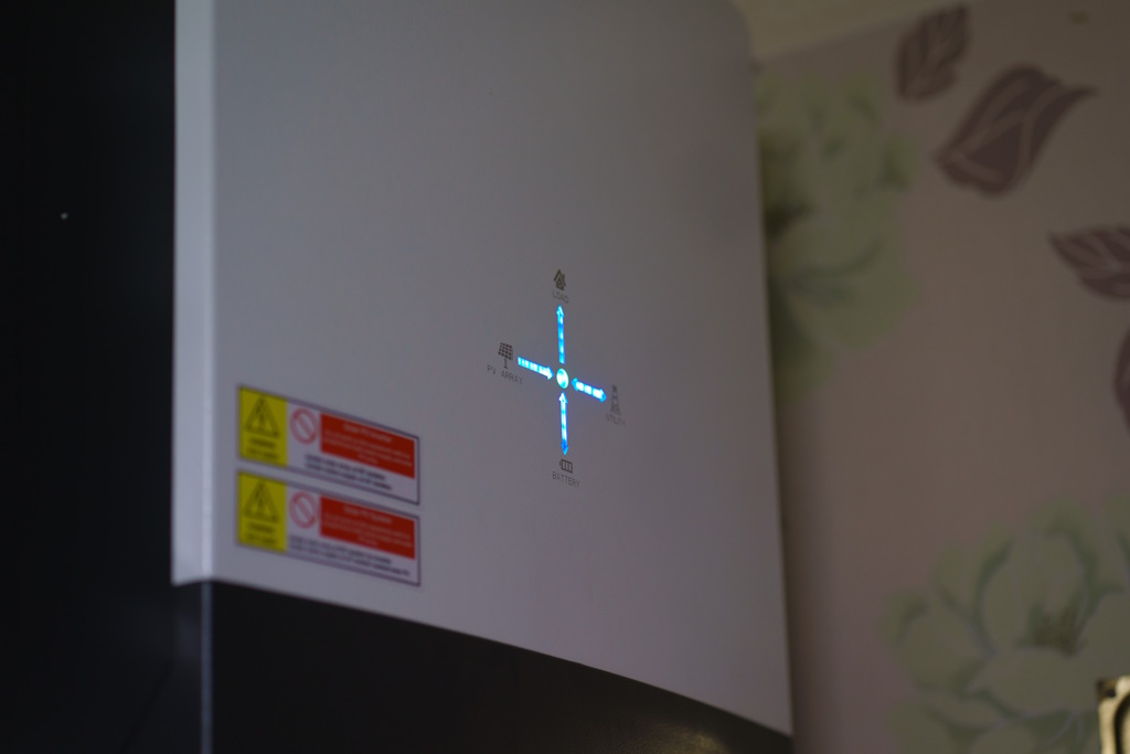 Solar panel inverter on a wall, with a white case and blue lights in the rough shape of a plus sign, against a white wallpaper