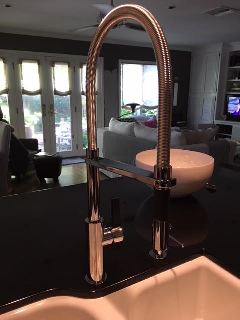 "A close-up view of a modern, polished chrome kitchen faucet with a tall, flexible, arched spout on a dark countertop. In the background, a well-lit, cozy living area is visible, featuring a large white bowl on a table, a couch, French doors, and a television.