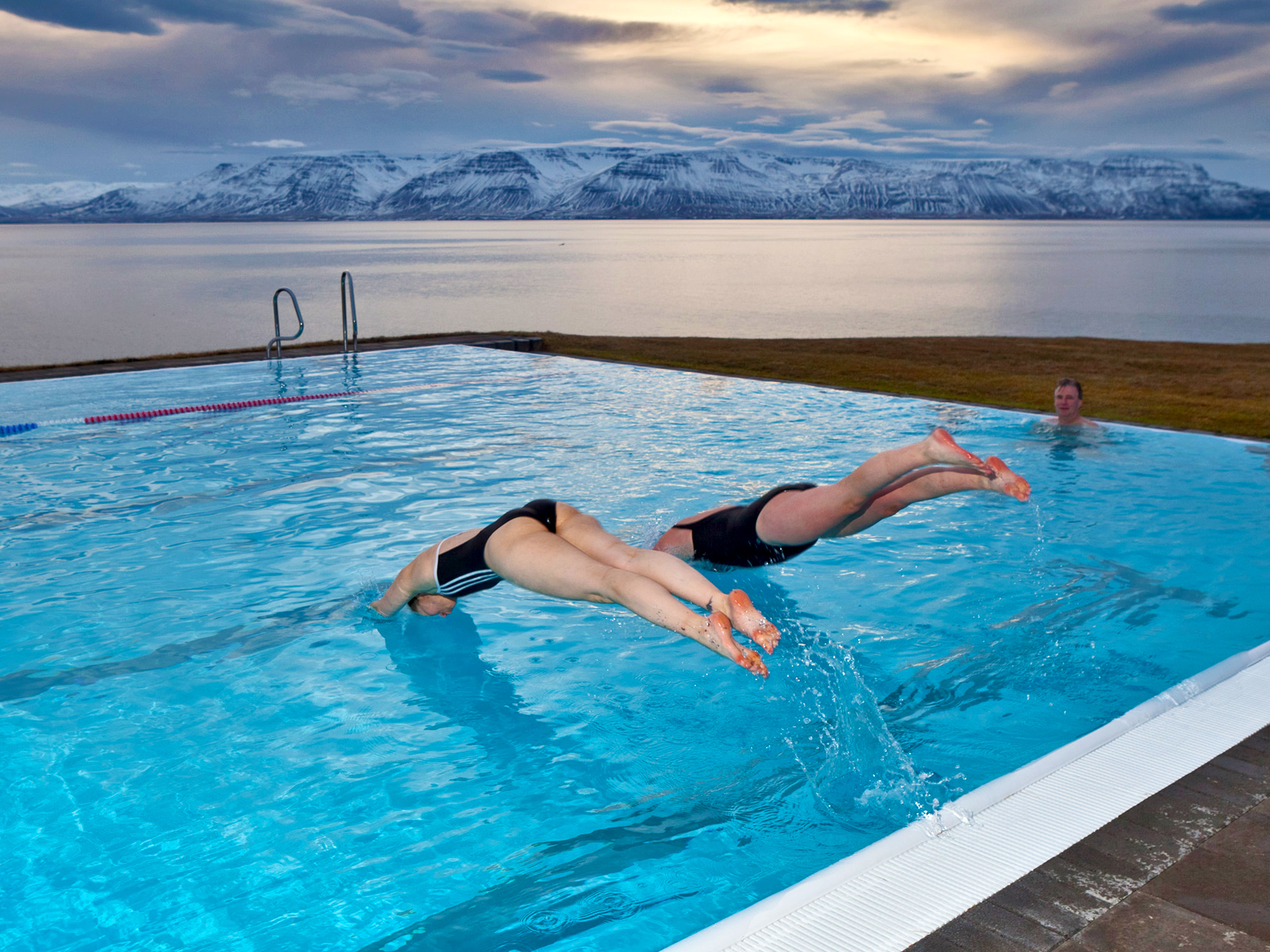 Two people diving into the Hofsós swimming pool with a fjord and mountains as a backdrop