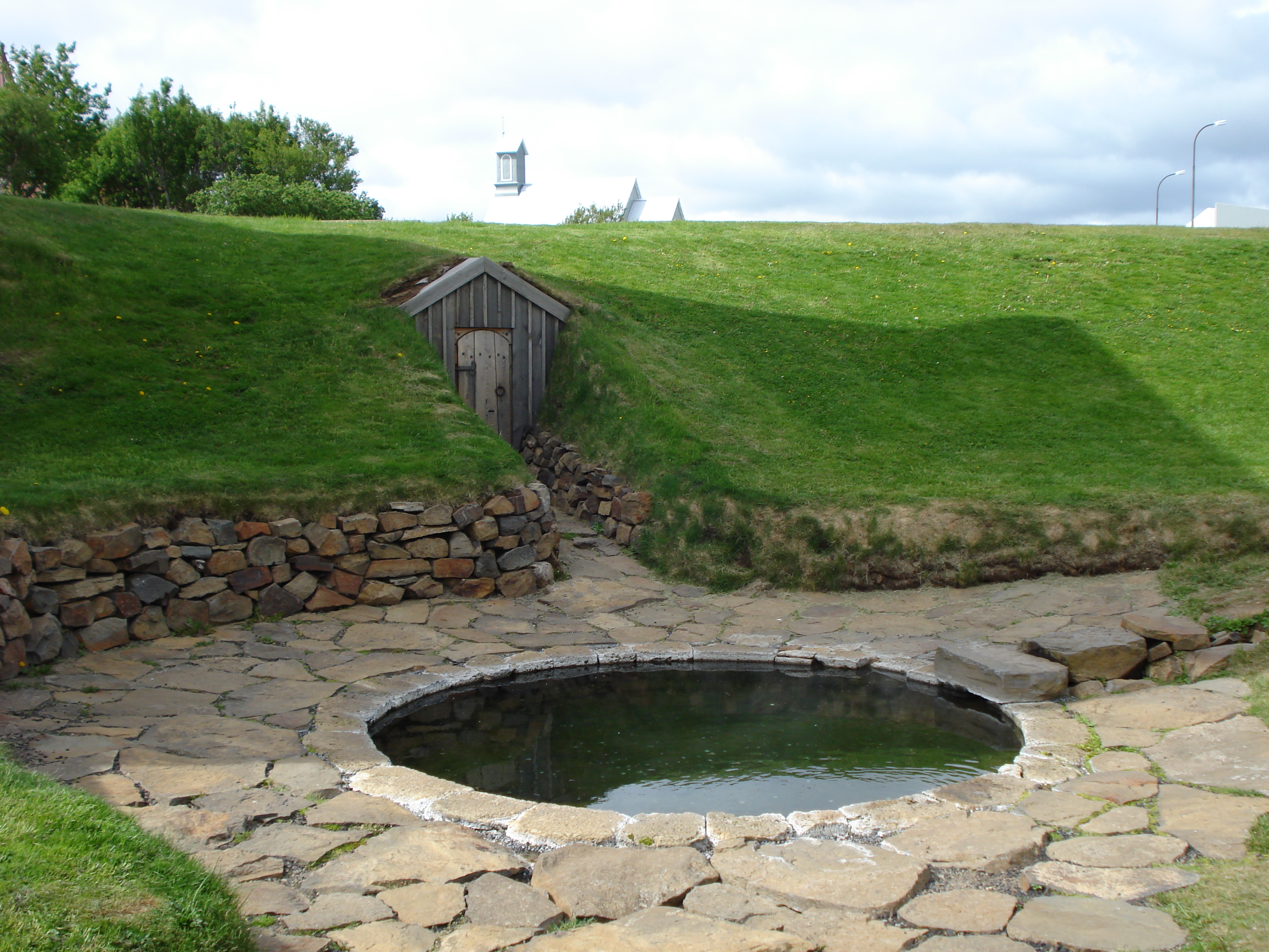 A round pool made of rocks in front of a sod house.