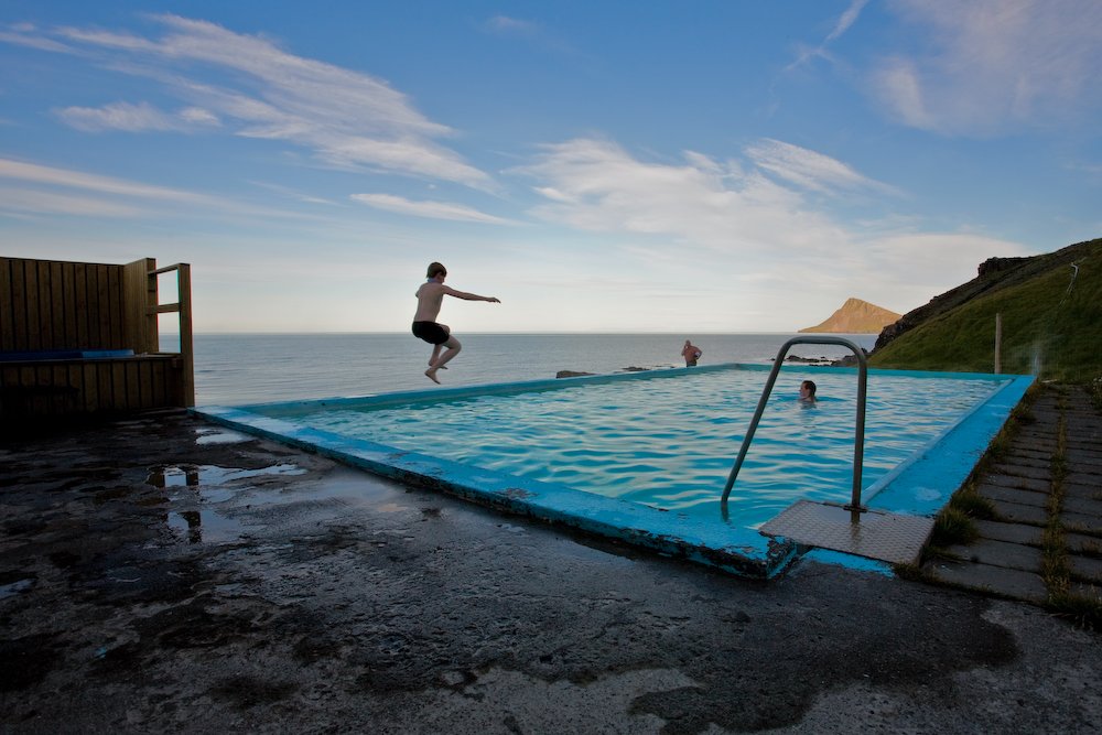 Young boy jumping into the pool at Krossneslaug, view of the mountains and the ocean in the background