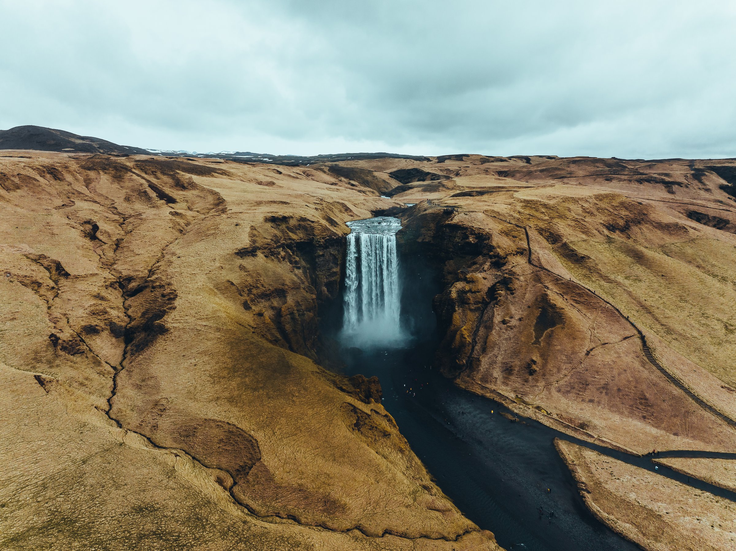 An aerial picture of Skogafoss in South Iceland. The landscape around the waterfalls is covered with brown grass, the water appears almost black.