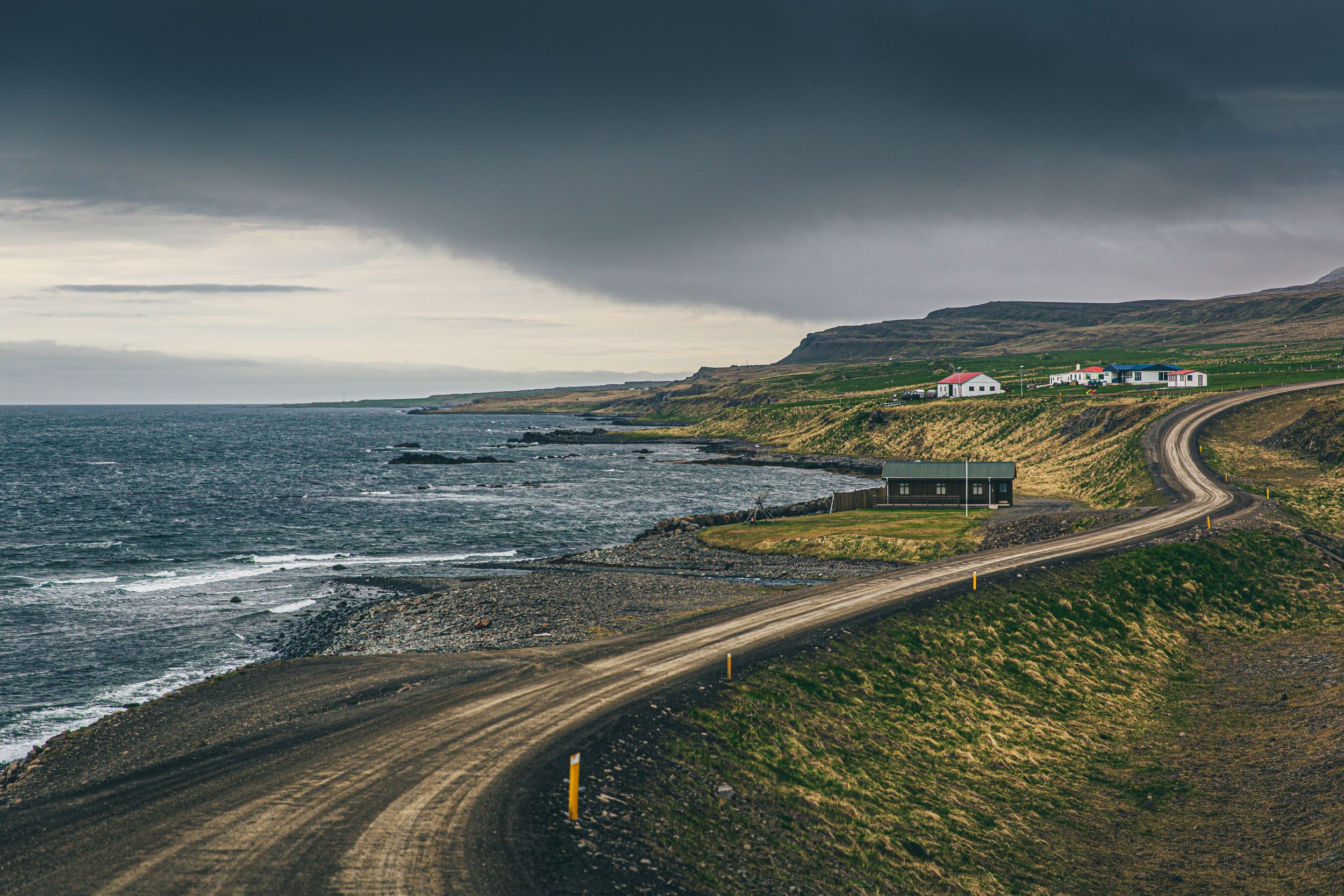 A curvy gravel road next to the ocean, farm houses in the background, cloudy skies.