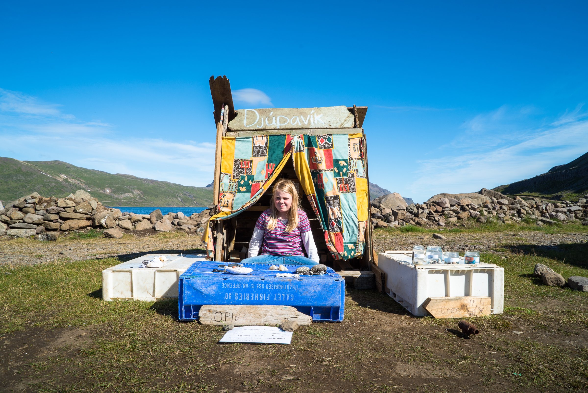 Young girl sitting in front of a self-made sales booth with the sign "Djúpavík" on it