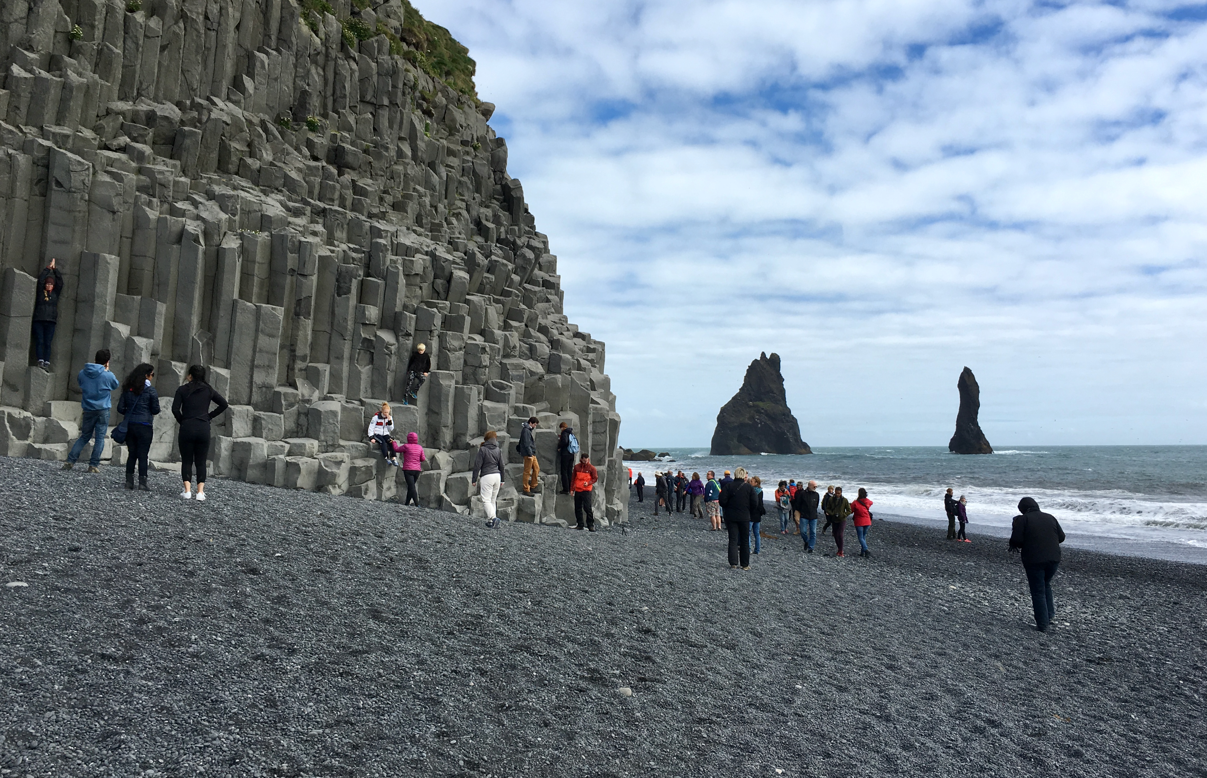 People standing on a black sand beach and climbing columnar basalt stacks.