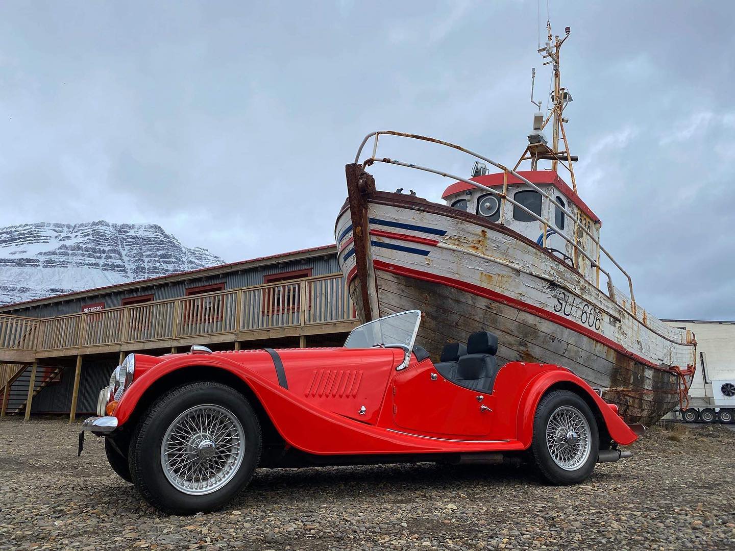 A bright red vintage car in front of an old boat