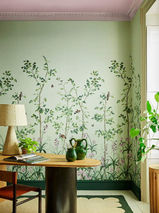 Green floral mural wallpaper featuring birds and butterflies (Bird & Bluebell - Pea Green) with a lamp and plant on a desk.